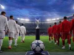 Dos equipos entran al campo, con un trofeo y un balón en primer plano